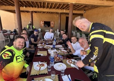 Grupo de participantes da Pegadas na Aventura a almoçar em Merzouga durante uma viagem de moto pelo deserto do Sahara, ambiente descontraído e companheirismo.