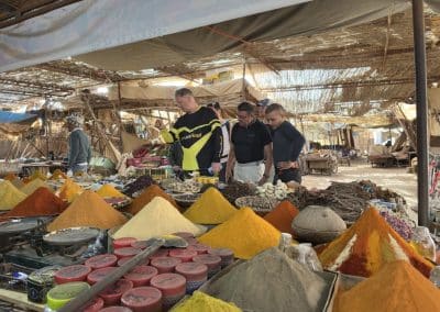 Motociclistas Pegadas na Aventura a explorar um mercado tradicional marroquino, com bancas de especiarias coloridas em Merzouga, Marrocos.