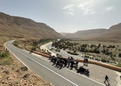 Grupo de motos da Pegadas n’Aventura a atravessar um vale no sul de Marrocos, numa estrada panorâmica entre montanhas, durante a viagem Al Maghrib 2026. Paisagens autênticas, cultura berbere e rota ideal para viagens de aventura em moto
