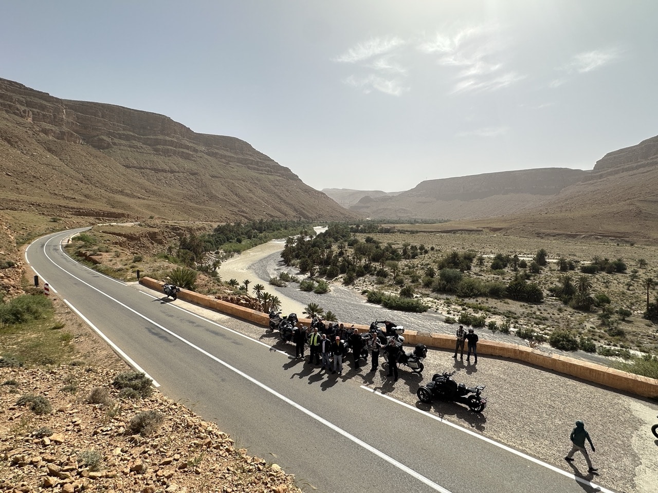 Grupo de motos da Pegadas n’Aventura a atravessar um vale no sul de Marrocos, numa estrada panorâmica entre montanhas, durante a viagem Al Maghrib 2026. Paisagens autênticas, cultura berbere e rota ideal para viagens de aventura em moto